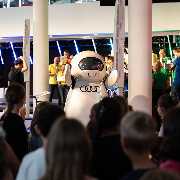 Kinder stehen vor einer Bühne mit einer Roboterfigur und Musikern bei einem Event der Audi Sommerkonzerte.