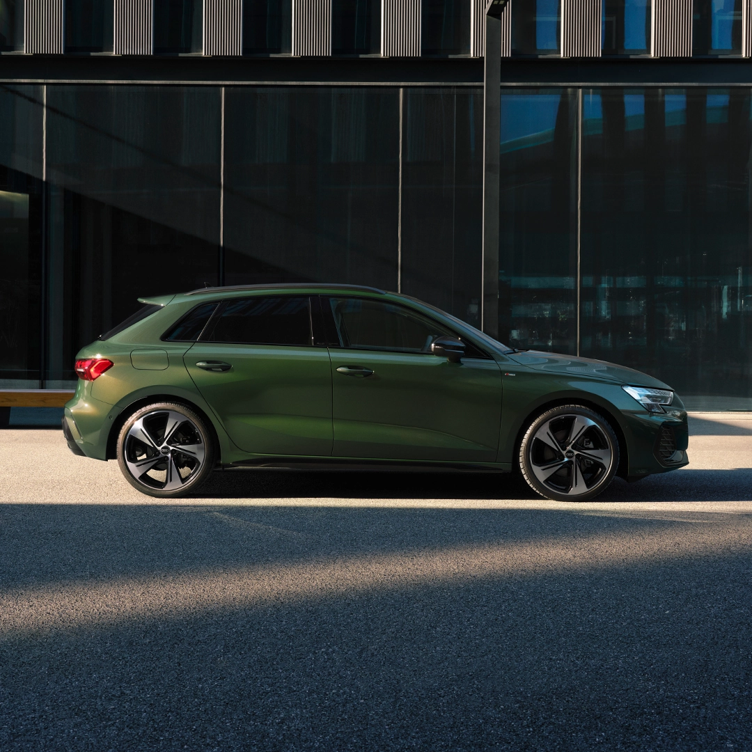 Green Audi A3 Sportback parked on road in front of building with large windows.