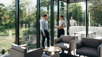 Zwei Personen stehen in einem modernen, verglasten Raum mit Blick ins Grüne und unterhalten sich bei Tageslicht.