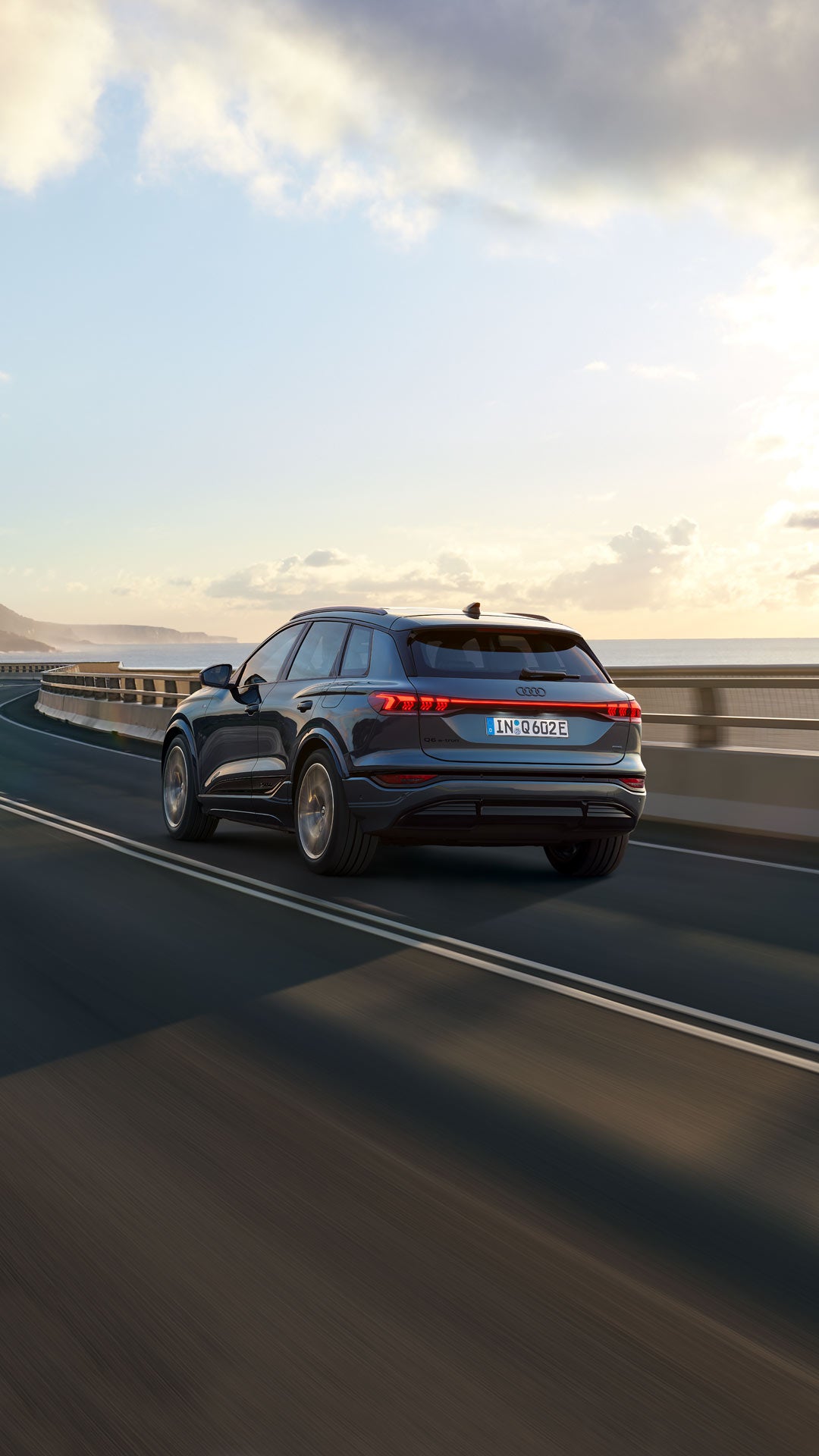 Rear view of the Q6 SUV e-tron driving along a winding road by the sea. Mountains can be seen in the background.