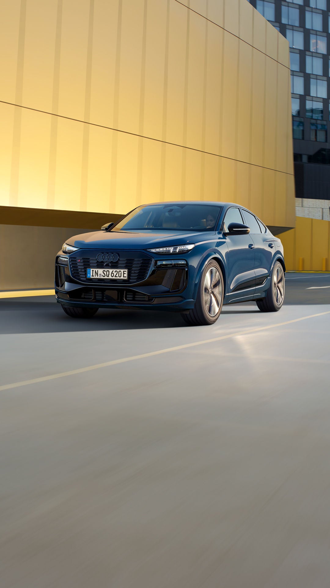 Schräges Frontprofil des SQ6 Sportback e-tron, welcher auf einer Straße durch die Stadt fährt. Im Hintergrund sieht man eine gelbe Gebäudefassade.