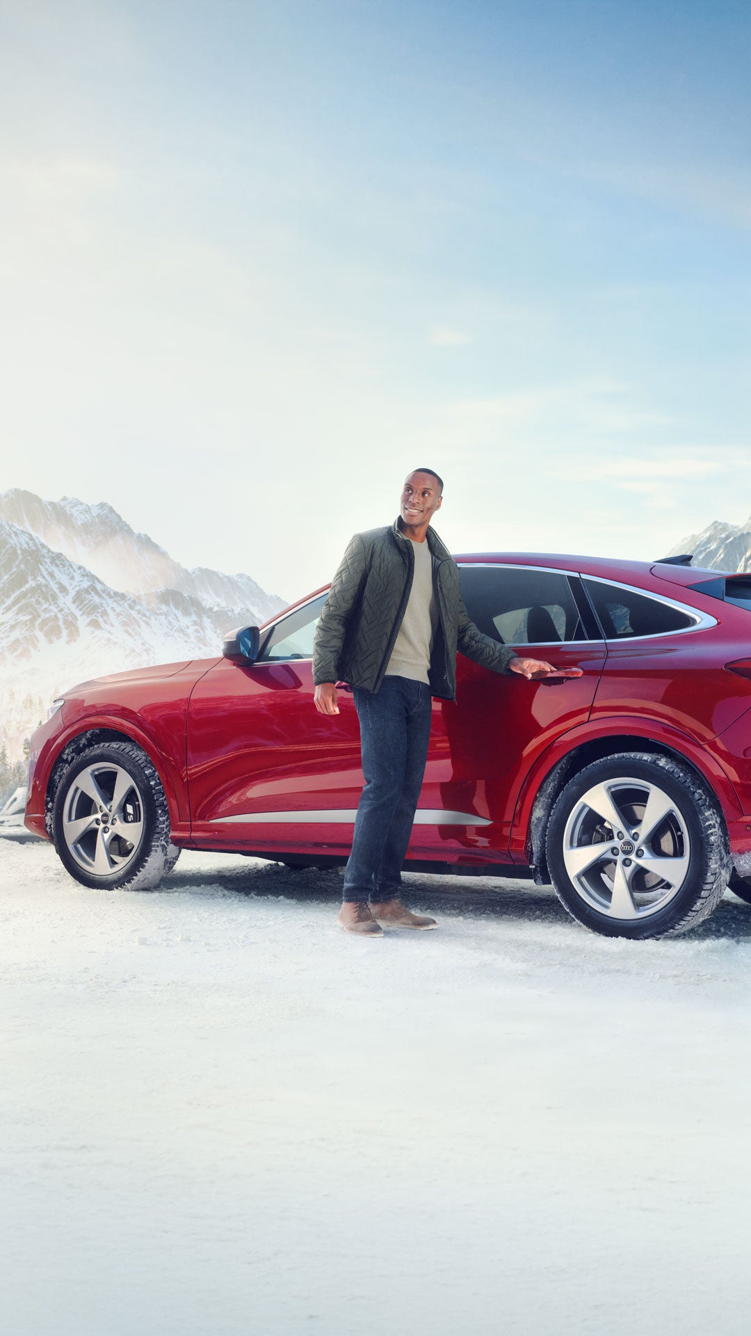 Person steht lächelnd neben einem roten SUV von Audi in verschneiter Berglandschaft.