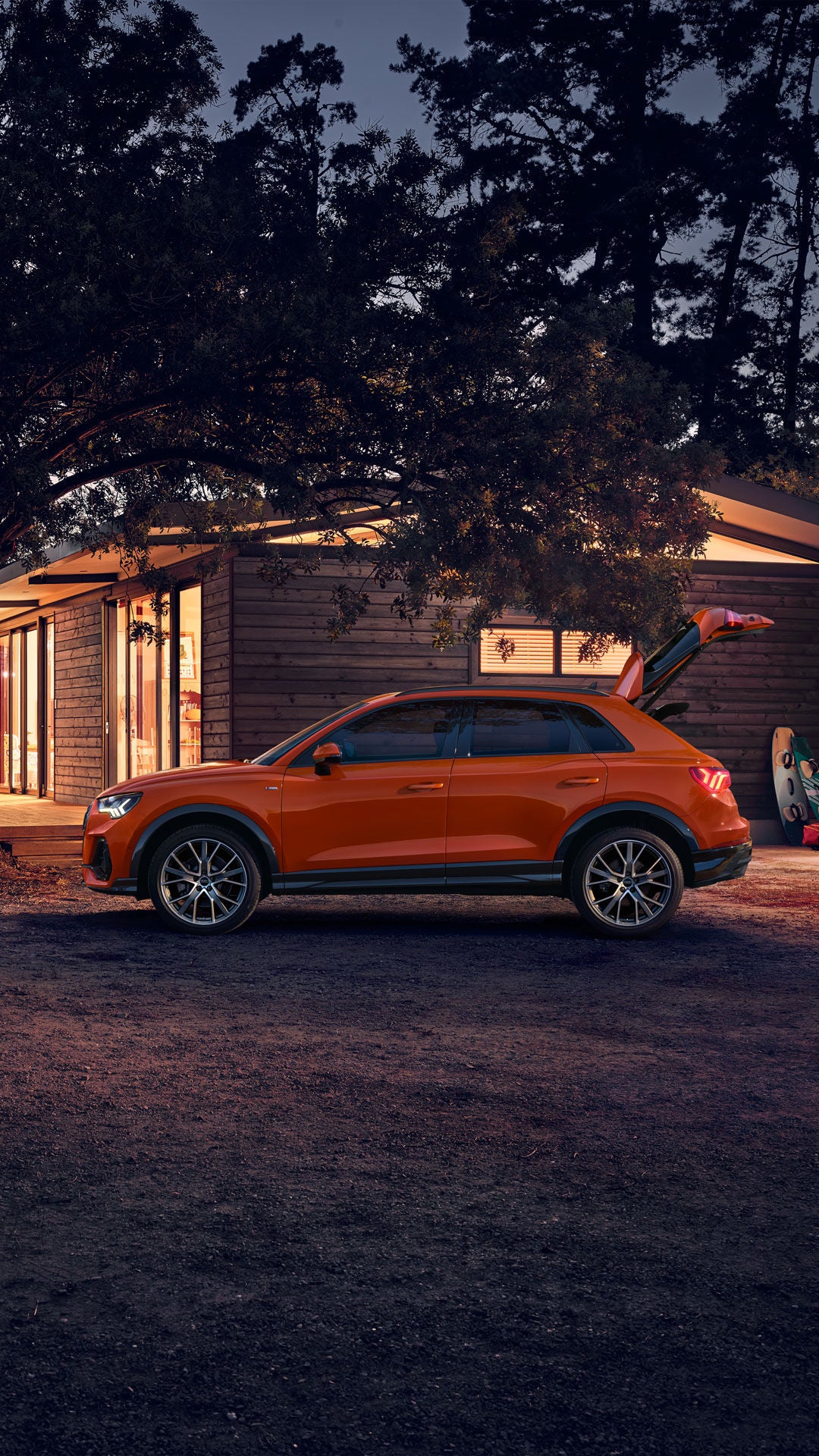 Ein orangefarbener SUV steht bei Dämmerung vor einem beleuchteten Holzhaus, umgeben von Bäumen und Freizeitgeräten.
