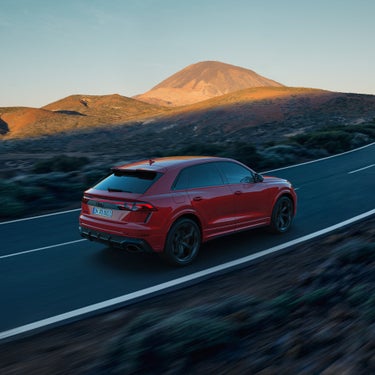 Ein roter Audi RS Q8 SUV performance fährt auf kurviger Landstraße durch hügelige Landschaft mit Berg im Sonnenuntergang im Hintergrund.