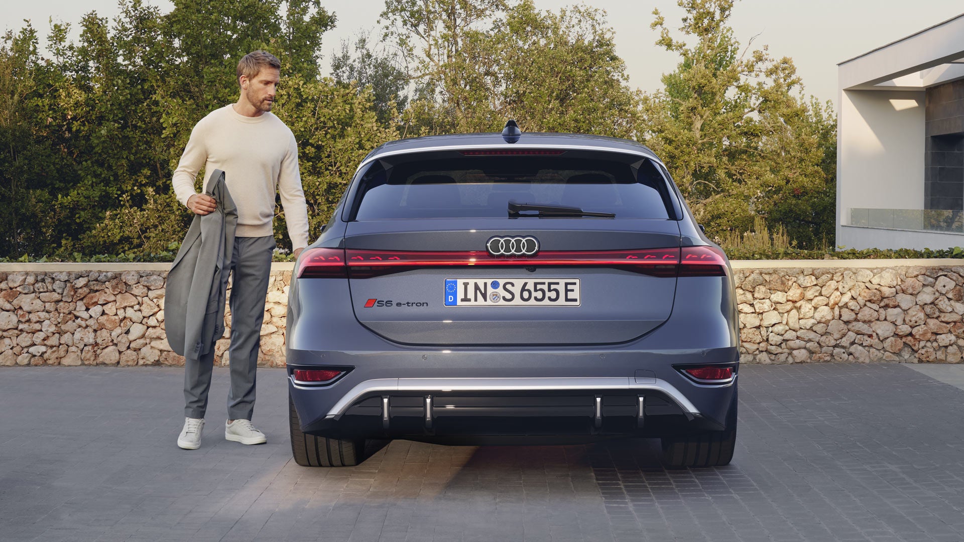 Heckprofil des S6 Avant e-tron, welcher auf Pflastersteinen vor einer Steinmauer und dem Garten eines modernen Hauses steht. Ein Mann mittleren Alters ist in Begriff, eine Tür zu öffnen.