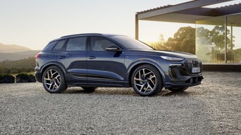 The Q6 SUV e-tron stands sideways on the gravel area in front of a modern house, illuminated by the setting sun.