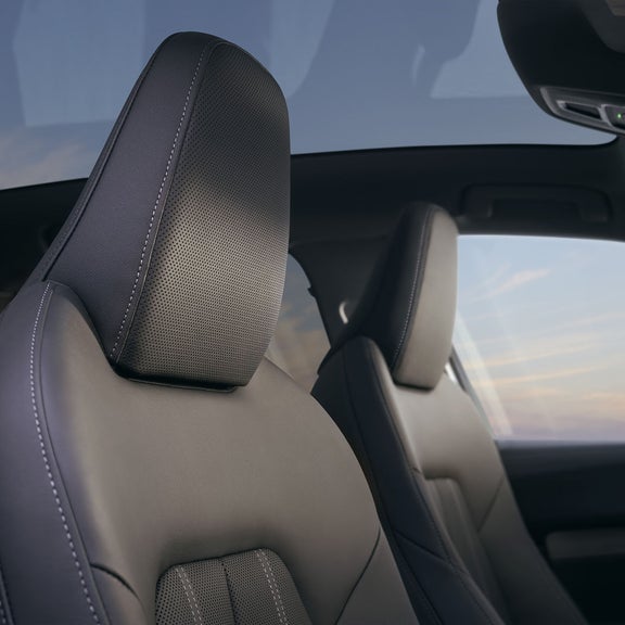 Close-up of a car's front seats upholstered in dark perforated leather with white stitching, set against a panoramic sunroof showing a blue sky.
