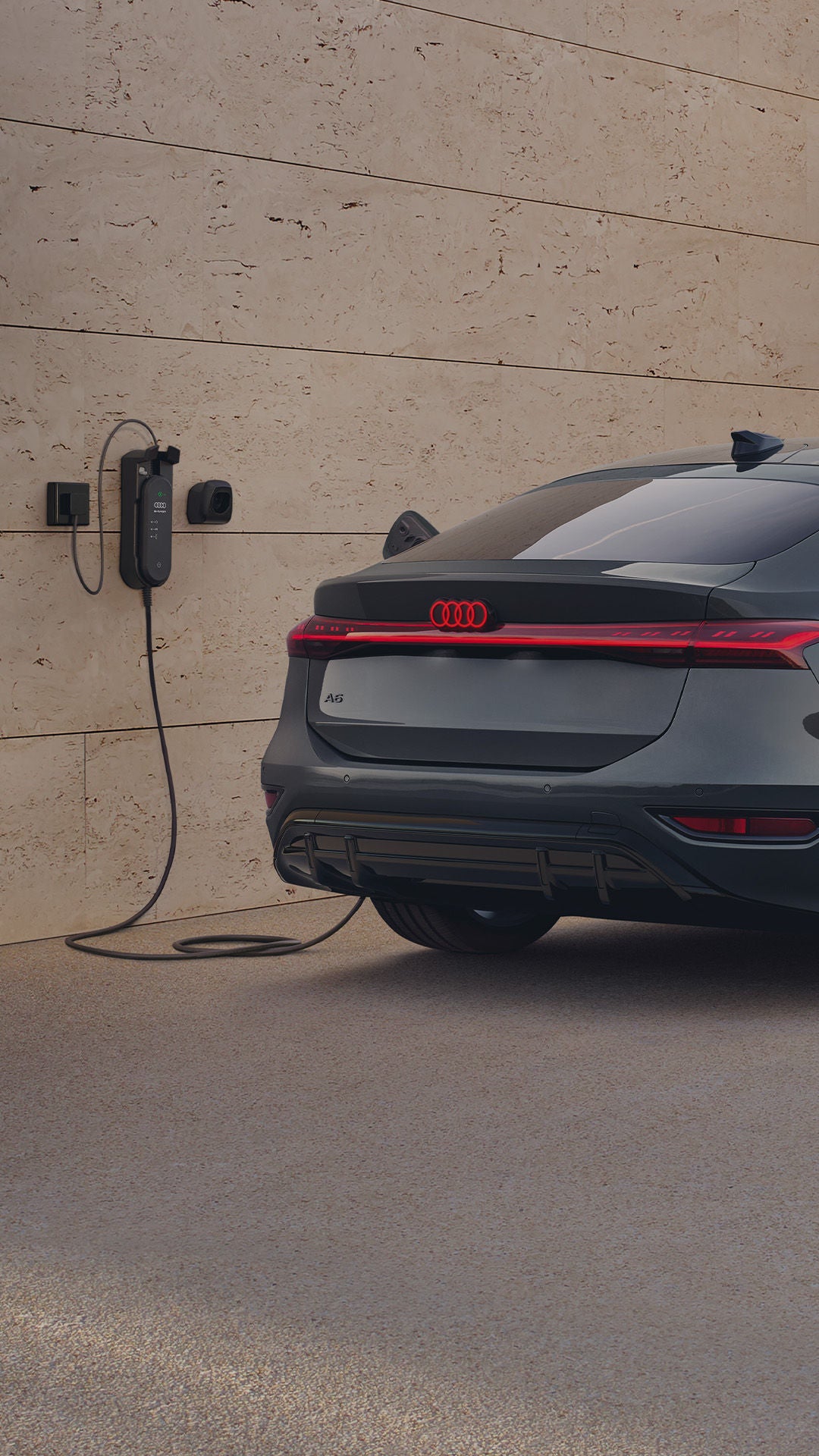 A grey Audi parked and charging at a wall-mounted electric charger next to a modern stone wall.