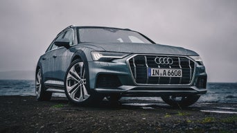 Grauer Audi auf nasser Straße am Meer bei bewölktem Himmel.