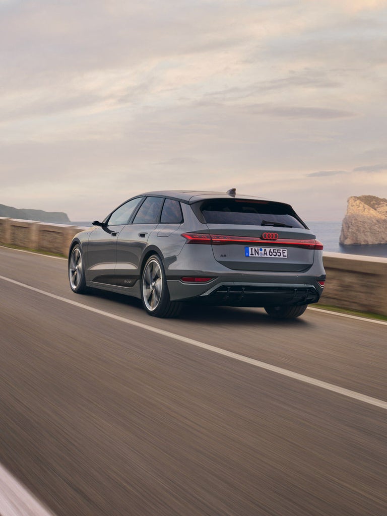 Grauer Audi A6 Avant e-tron fährt auf Küstenstraße, Meer und Felsen im Hintergrund.
