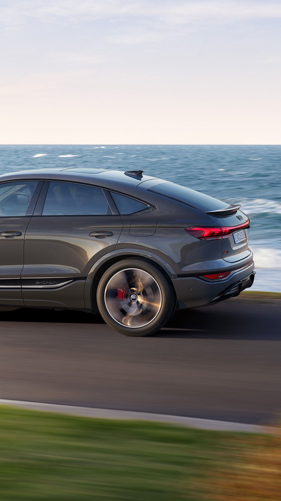 A sleek grey Q6 Sportback e-tron speeds along a coastal road, with waves crashing nearby and lush green hills in the background.