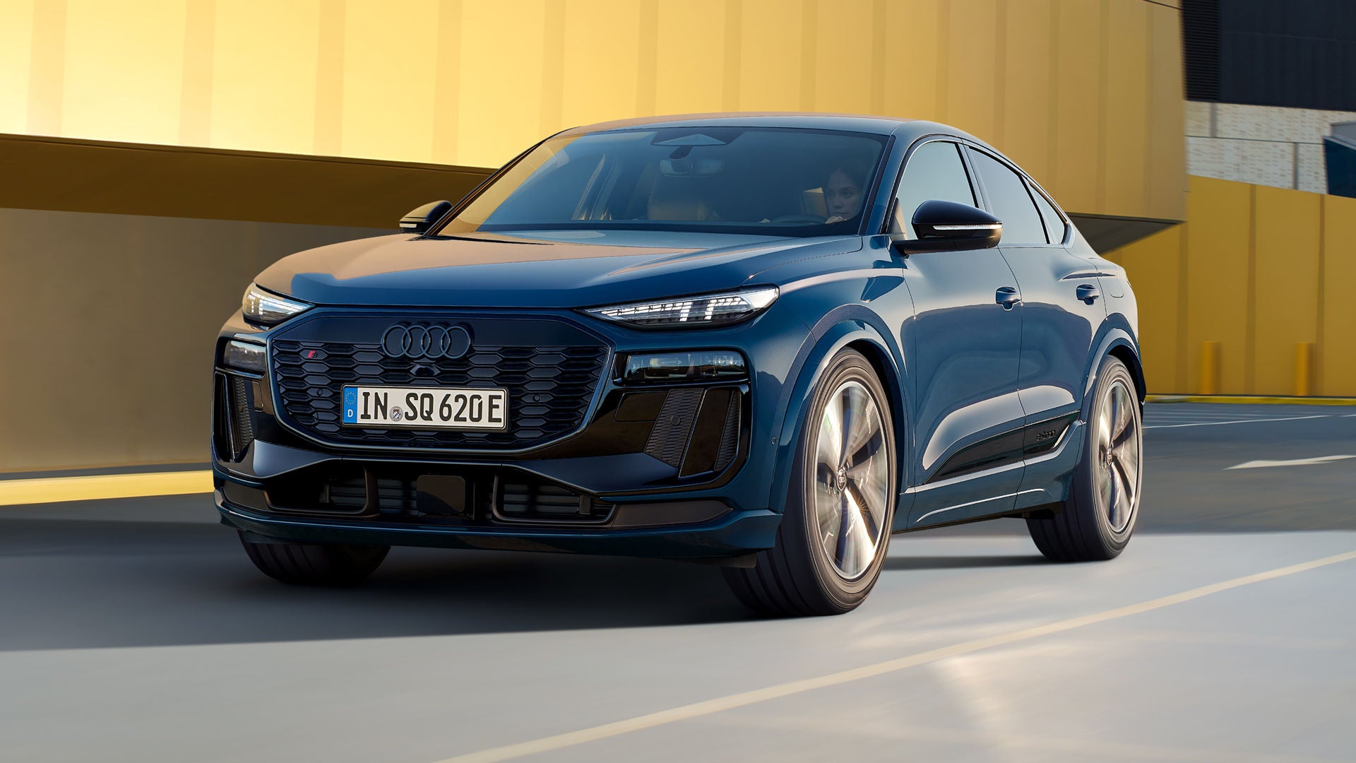 Schräges Frontprofil des SQ6 Sportback e-tron, welcher auf einer Straße durch die Stadt fährt. Im Hintergrund sieht man eine gelbe Gebäudefassade.