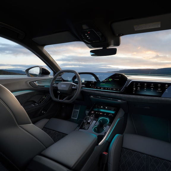 Car interior with sleek design, digital dashboard displaying controls, and a scenic view of a lake and mountains at sunset through the window.