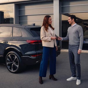 A woman in a beige jacket receives car keys from a man in a grey sweater and white shirt. They are standing near a dark SUV with a modern building behind them.
