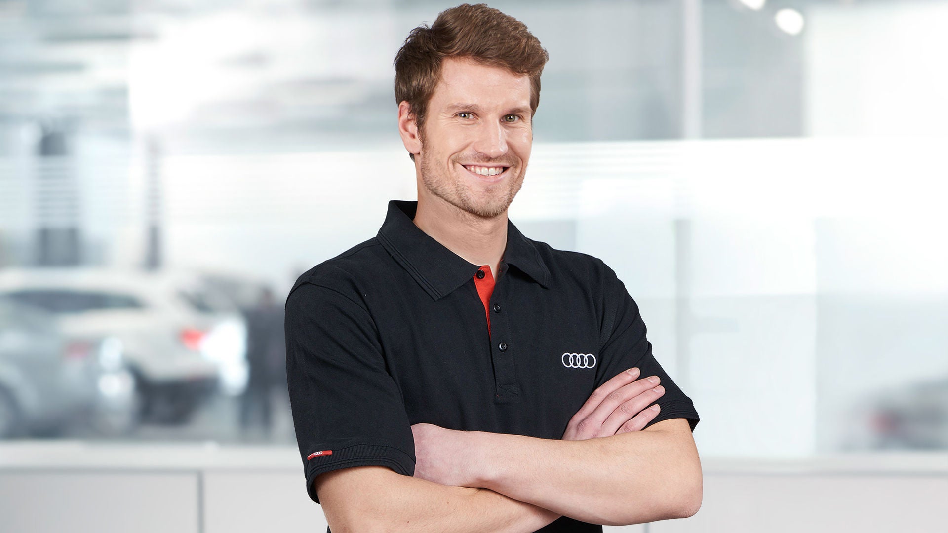 Kfz-Servicetechniker in einem schwarzen Poloshirt steht mit verschränkten Armen im Autohaus und lächelt in die Kamera.