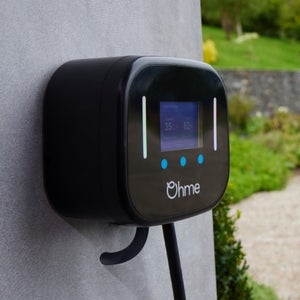 A black Ohme electric vehicle charger mounted on a concrete wall, displaying charging status. It's set outdoors, with a garden and path visible in the background.