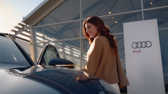 Une femme rousse vêtue d'un manteau marron se tient devant un concessionnaire Audi et monte dans une voiture.