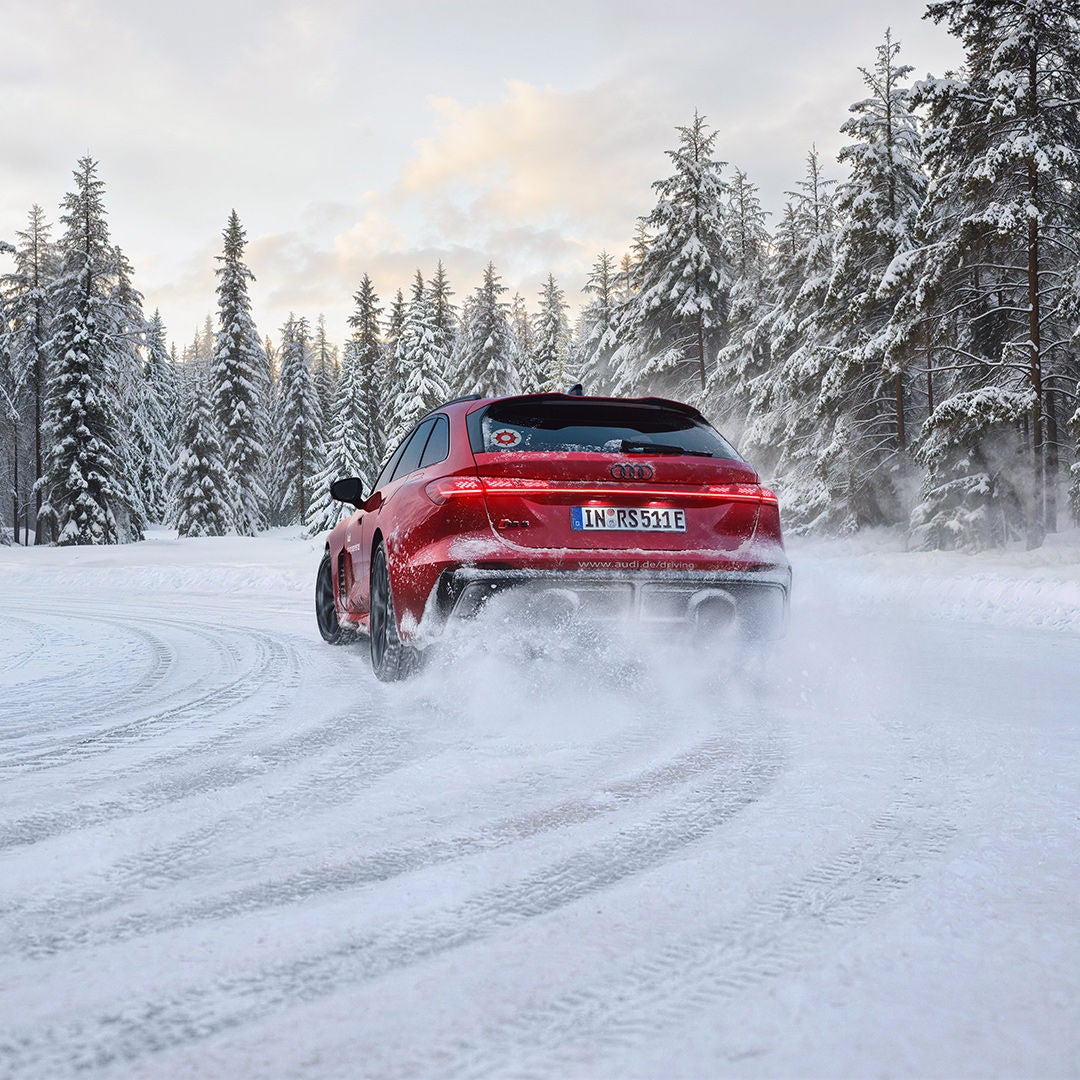 Rear view of a red Audi RS 5 Avant on a snow-covered road; swirling snow, tire tracks, and a forest.
