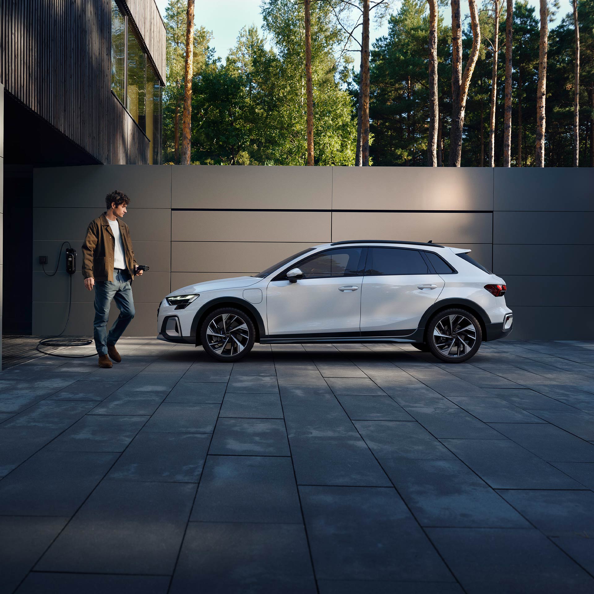 Ein Mann mit Ladekabel steht vor einem weißen Audi A3 allstreet TFSI e, der vor einer modernen Wand geparkt ist.