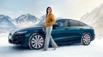 Frau in gelber Jacke steht neben blauem Audi auf verschneiter Fläche vor Bergkulisse.