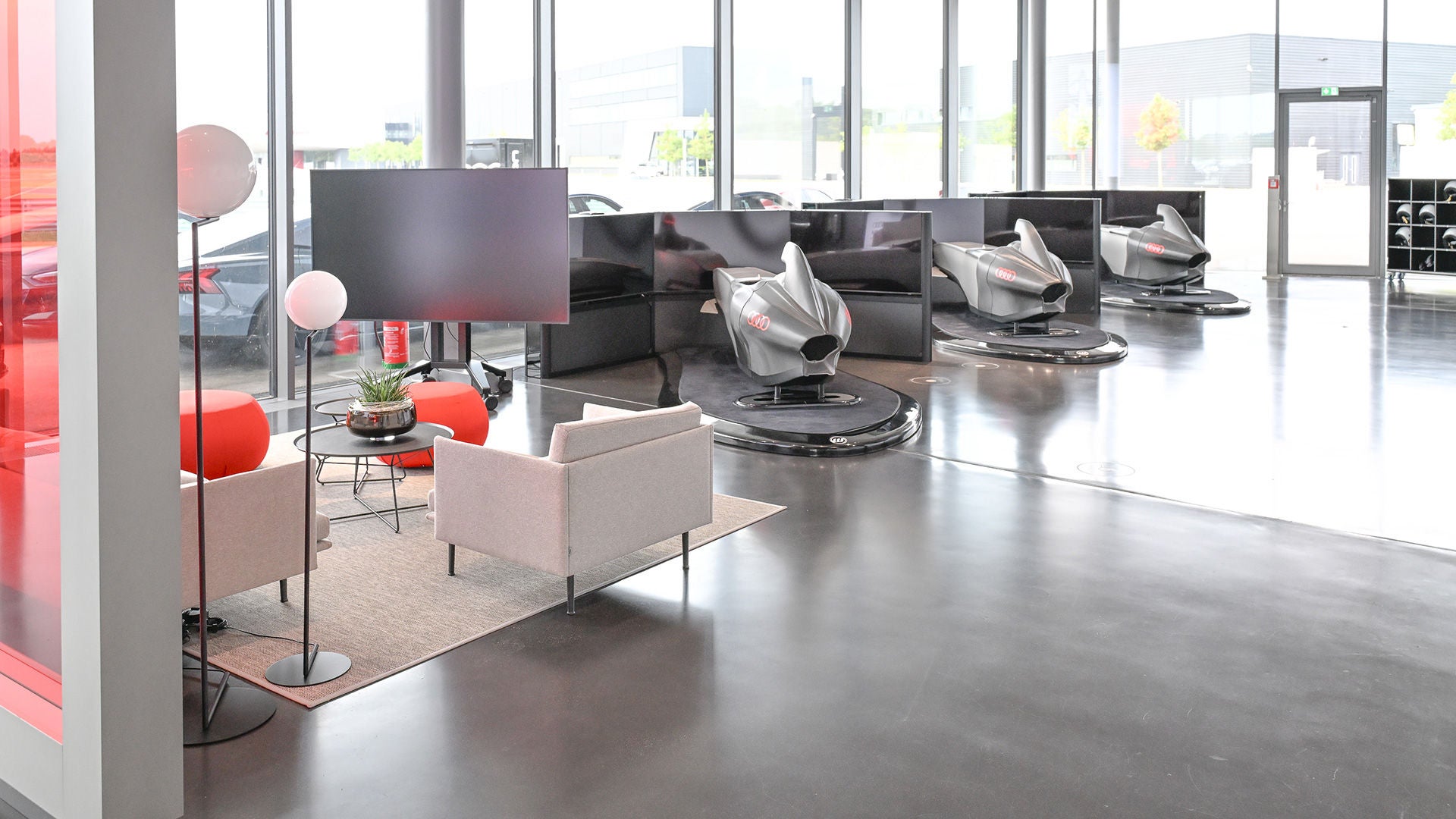 Heller Showroom mit drei silbernen Audi-Rennsimulatoren und einer Lounge mit hellen Sesseln und roten Akzenten.