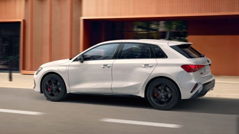 A white Audi A3 Sportback car driving on a city street, side view. Car has sleek design and black rims. Background shows orange building facade, blurred.