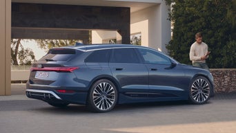 Der S6 Avant e-tron steht schräg seitlich auf Pflastersteinen vor einem modernen Gebäude. An einer am Gebäude befestigten Steinmauer hinter dem Fahrzeug ist ein Mann mittleren Alters gelehnt.
