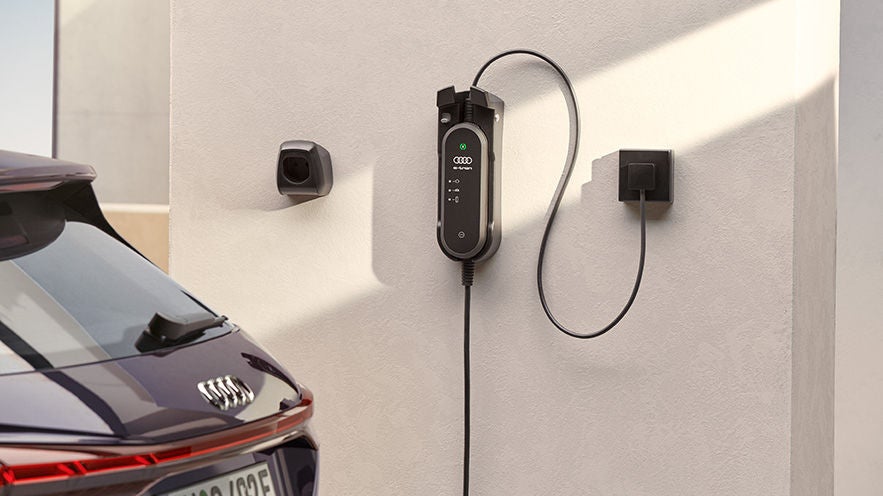 Heimladestation mit Audi Ladegerät an einer Wand, daneben das Heck eines parkenden Elektrofahrzeugs.