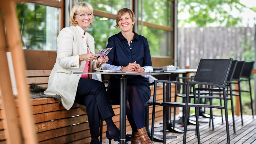 Zwei Frauen (Gudrun Sander und Antonia Wadé) sitzen an einem Tisch im Freien, eine hält Prospekte, beide lächeln in die Kamera.