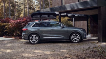Ein grauer Audi Q5 mit Dachbox steht unter einem Carport. Im Hintergrund ist ein Wald zu sehen.