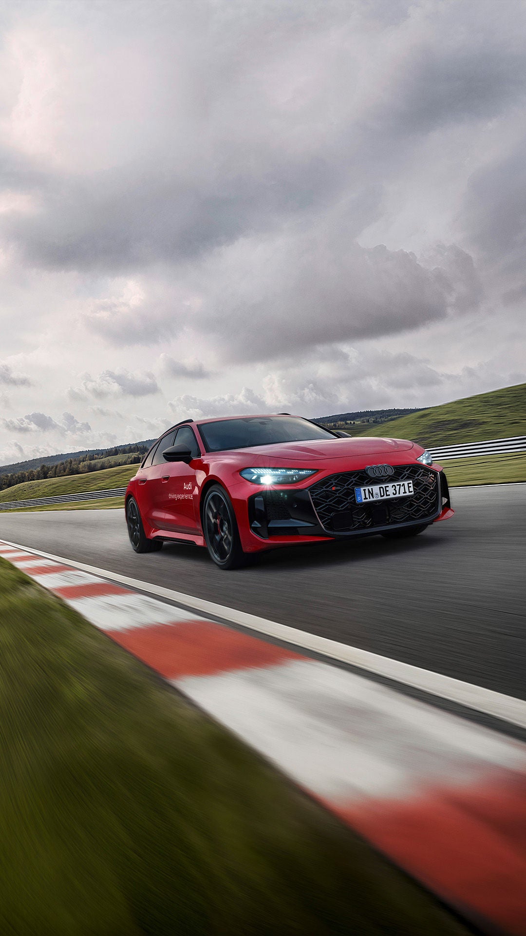 Roter Audi RS 5 Avant auf Rennstrecke in Kurve, dynamische Perspektive mit Leitplanke und bewölktem Himmel.