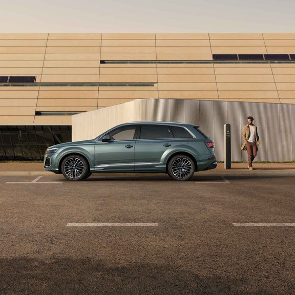 Der grüner Q7 SUV TSFI e parkt auf einer Seitenstraße vor einem modernen Gebäude. Daneben steht ein schlicht gekleideter Mann.