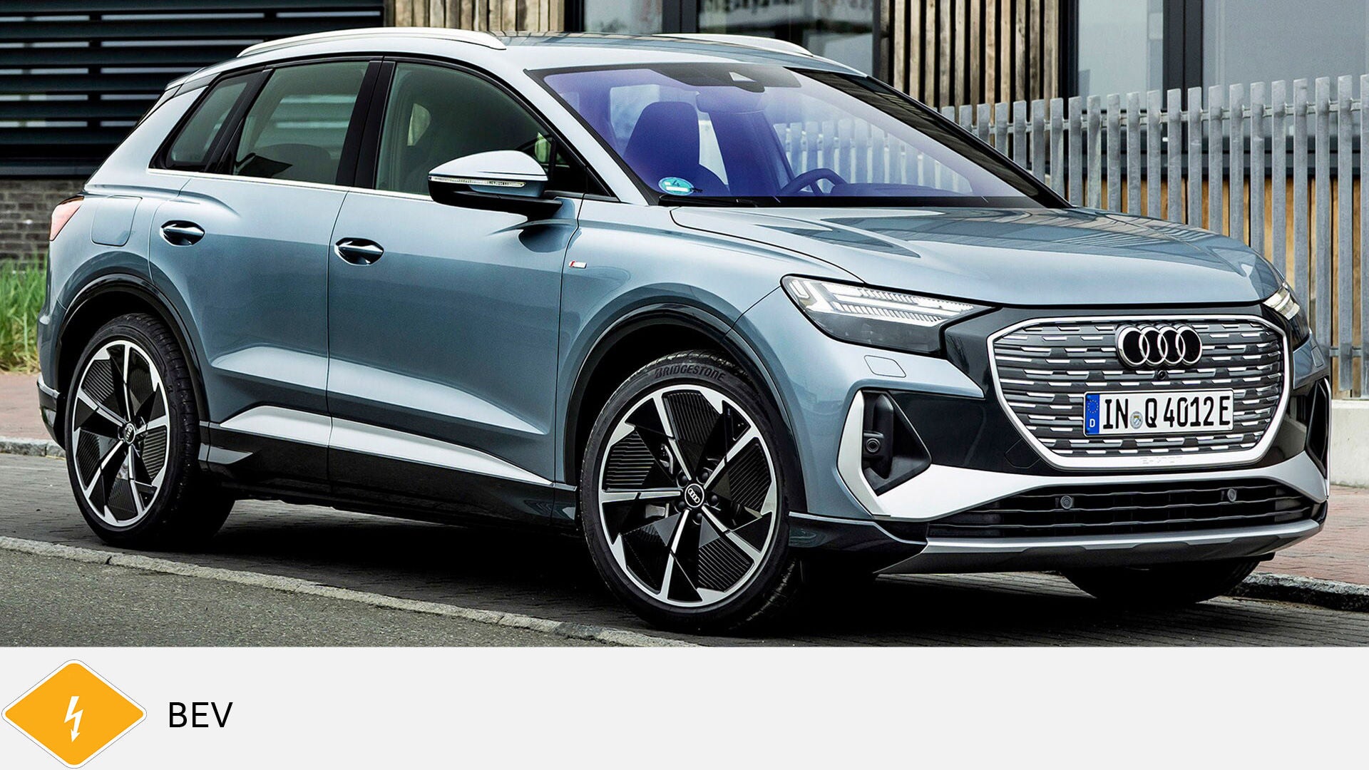 Gray Audi Q4 e-tron SUV standing in front of building with wood and metal facade. Yellow BEV symbol with lightning bolt below.