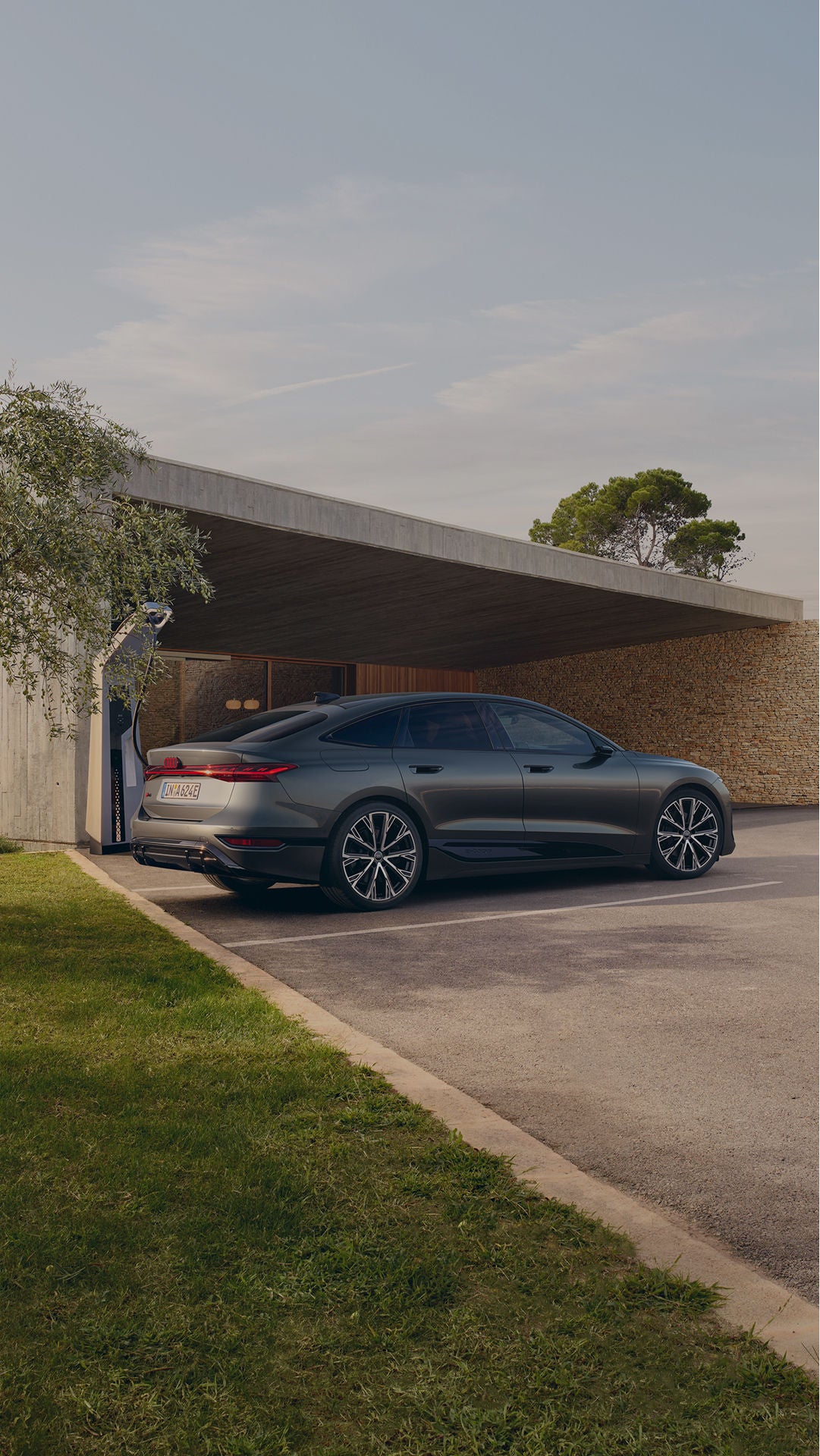 A sleek grey sedan parked outside a modern house under an open garage, near trees and a stone wall.