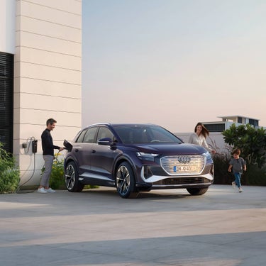 Mann lädt violetten Audi Q4 e-tron an Wallbox, während Frau mit Kind von einem modernen, hellen Haus angelaufen kommen.