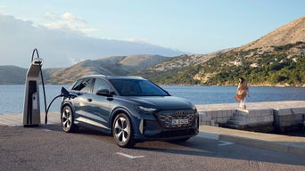 Audi Q4 SUV e-tron lädt an Ladestation am Meer, während eine Person am Ufer entlanggeht.