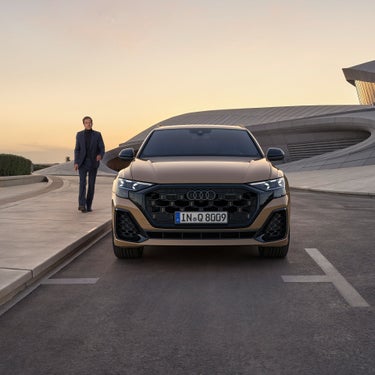 Ein goldener Audi Q8 SUV frontal auf einer Straße, links daneben ein Mann im Anzug.