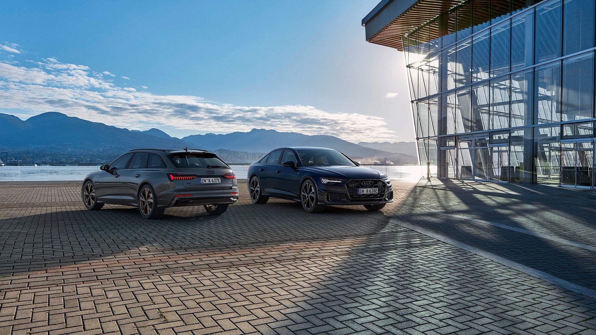 Zwei Autos stehen auf einem Parkplatz vor einer berigen Kulisse und Wasser, daneben ein moderne Haus aus Glas.