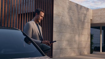Person hält Smartphone mit Fahrzeug-App in der Hand, im Hintergrund ein geparkter weißer Audi.