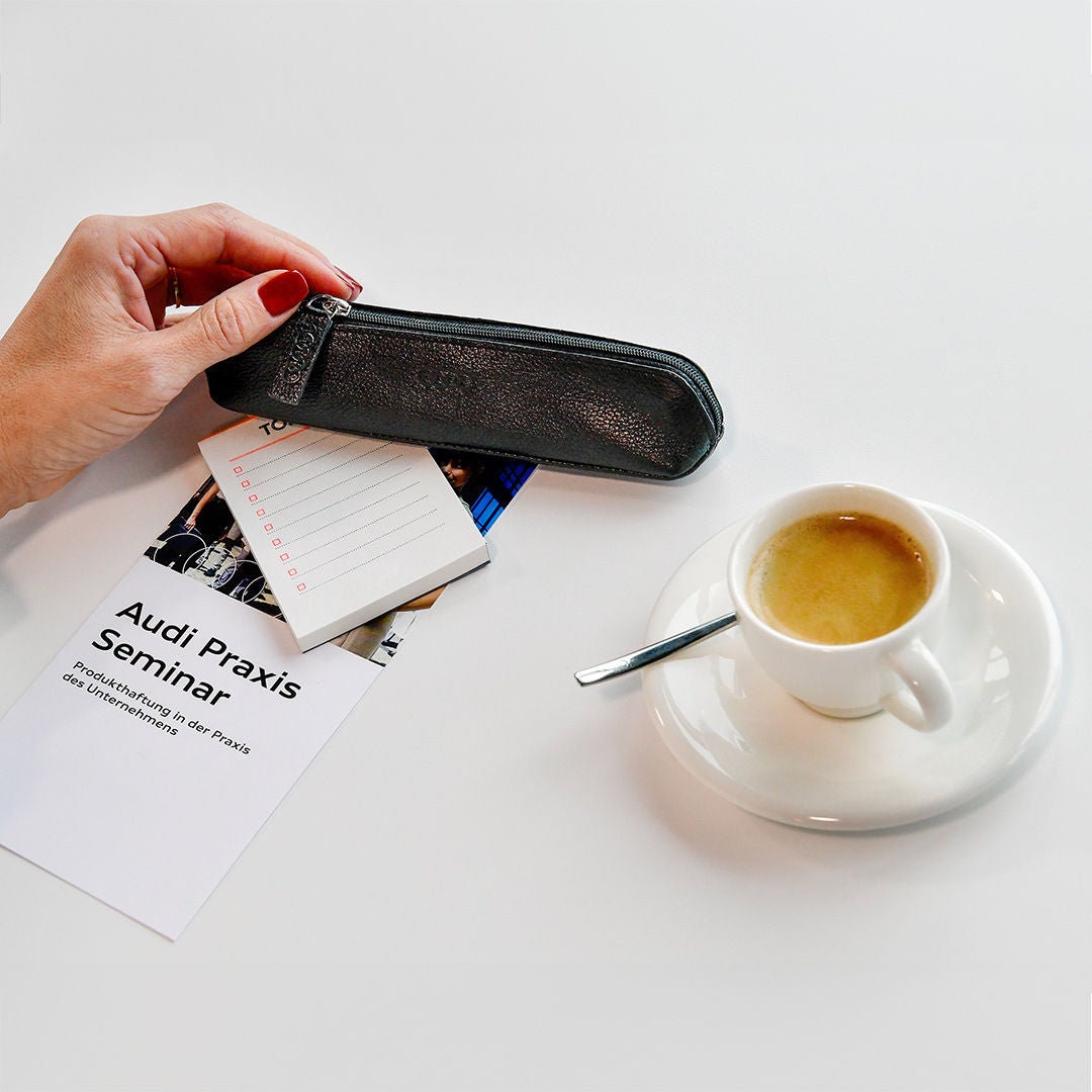 Hand mit rotem Nagellack hält ein schwarzes Etui neben Espresso, Notizblock und Audi-Seminarbroschüre auf weißem Tisch.