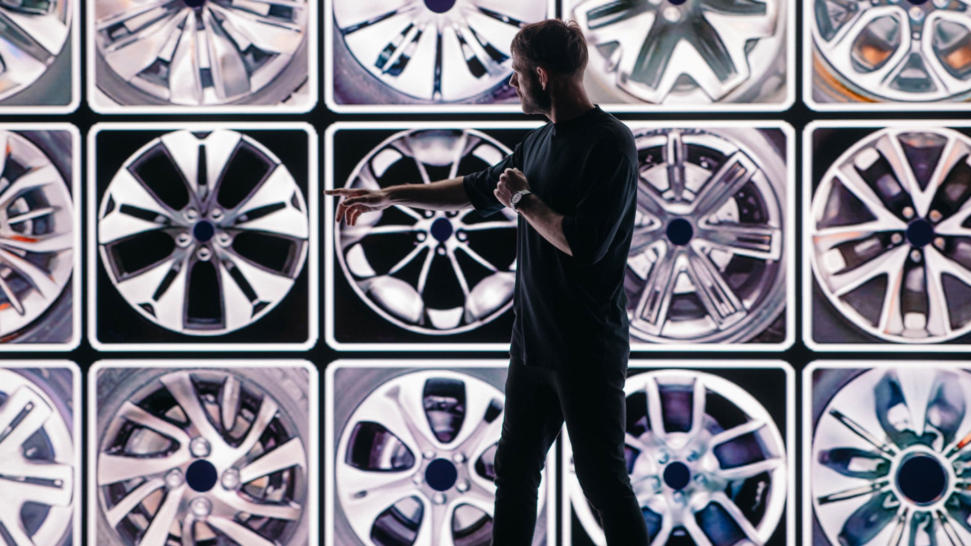 A man stands in front of a video wall displaying wheel rim designs and points to a motif.