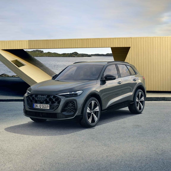 Der Q5 SUV in der edition one mit Kontrasten in Mythosschwarz steht im seitlichen Frontprofil auf einem Parkplatz direkt an der Küste. Dahinter ein futuristisches Haus.
