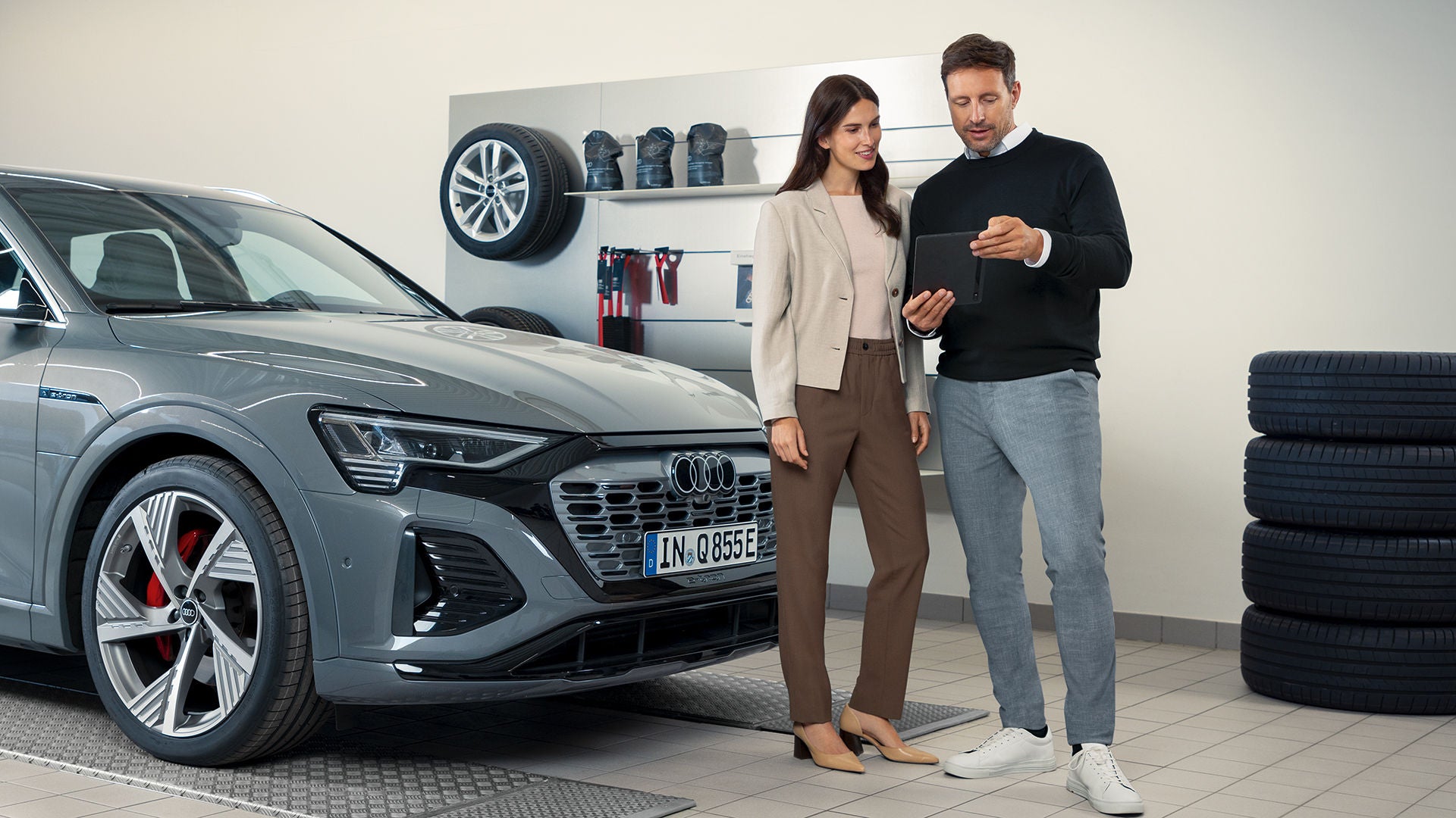 Ένας άντρας και μια γυναίκα στέκονται μπροστά από ένα γκρι Audi, κοιτάζοντας κάτι σε ένα iPad.