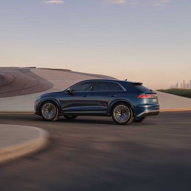 Ein dunkelblauer Audi SQ8 SUV fährt auf moderner Straße mit geschwungener Architektur im Hintergrund.