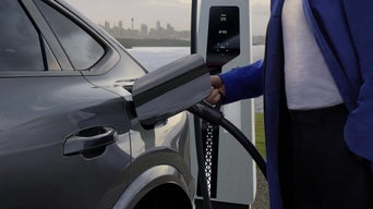 Close-up of the charging socket of the Q6 Sportback e-tron, into which a woman is plugging a charging cable. In the background is the sea and the skyline of a city.