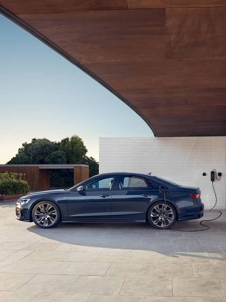 Dunkelblauer Audi A8 60 TFSI e lädt an Hauswand mit Ladesystem, moderne Umgebung mit Pflanzen im Hintergrund, Seitenansicht.