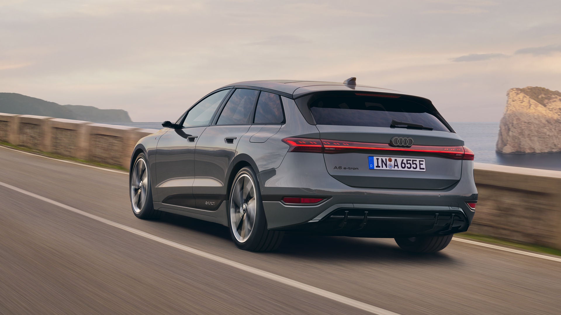 Schräge Heckaufnahme des A6 Avant e-tron, welcher auf einer Straße entlang des Meeres fährt. Im Hintergrund sind Felsen und Berge zu erkennen.