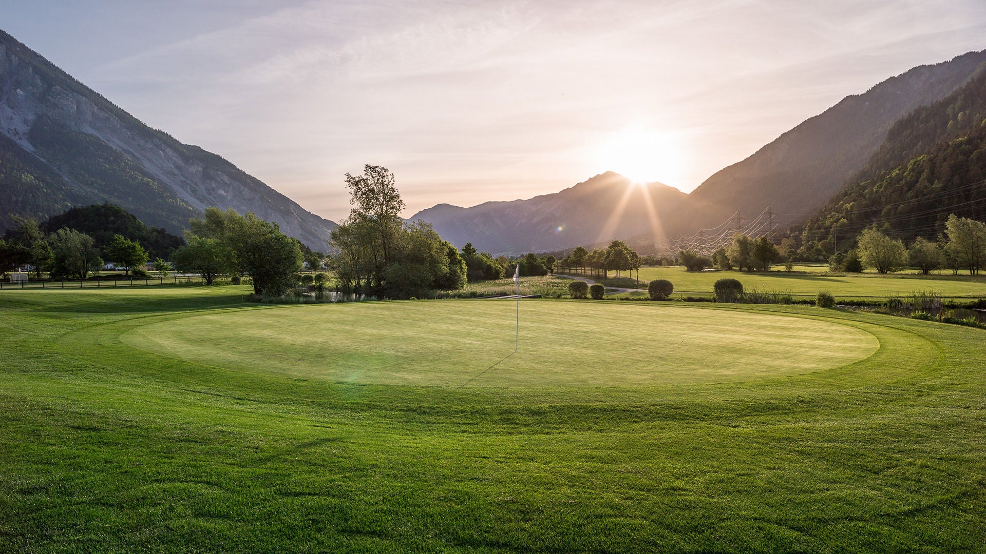 A golf green in a valley at sunset, surrounded by mountains and trees.