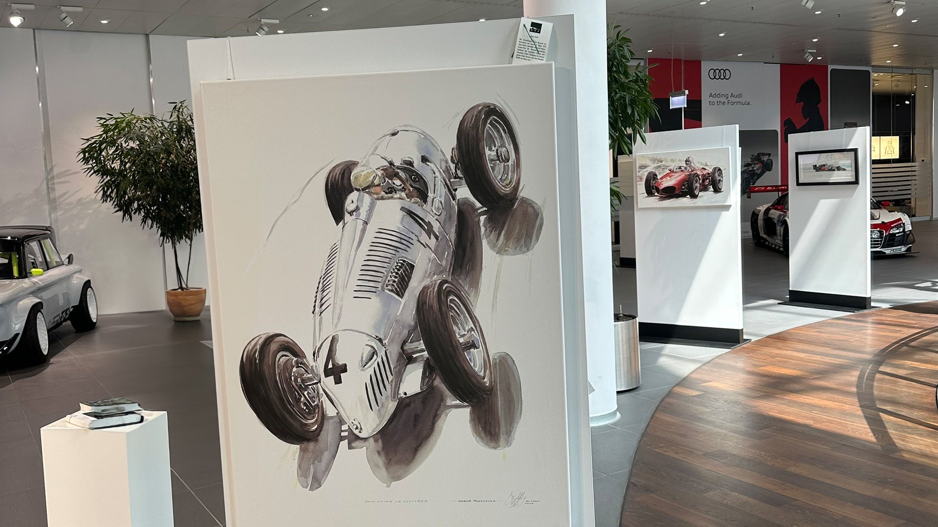 Gemälde eines silbernen Rennwagens mit Nummer 4 in Vogelperspektive, ausgestellt in einem modernen Audi-Showroom mit weiteren Kunstwerken.