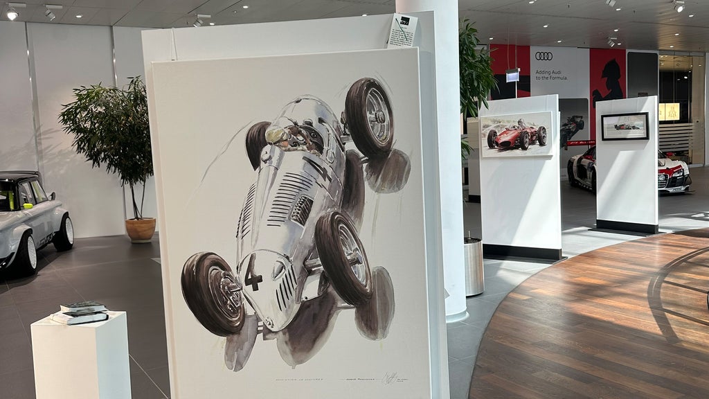 A painting of a silver race car bearing the number 4, viewed from a bird's-eye perspective, on display in a modern Audi showroom alongside other works of art.